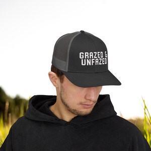 May include: A black and gray trucker hat with the text "GRAZED & UNFAZED" in white block letters. The hat has a mesh back and a curved brim. The hat is worn by a person wearing a black hoodie.