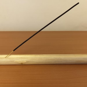 May include: A light-coloured wooden incense holder with a single, lit incense stick. The holder is a rectangular block with a groove. The incense stick is dark brown and angled upwards.
