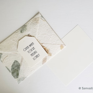 May include: A white envelope with a tag that says "Open when you're feeling lonely". The envelope is made of textured paper with brown and green floral accents.