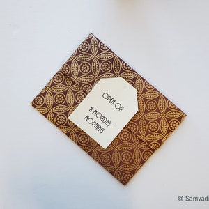 May include: A folded brown and gold patterned card with a white tag that reads "OPEN ON A MONDAY MORNING". The card has a repeating floral design. The tag is attached to the card. The background is white.