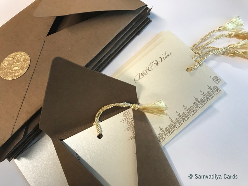 May include: A stack of brown envelopes with a gold seal on the top envelope. The envelopes are open to reveal a white card with a gold border and the text "Best Wishes". The card has a gold tassel attached to it.