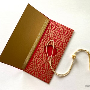 May include: Red envelope with a gold patterned design and a gold tassel attached to a hole in the envelope. The envelope is open and the inside is lined with gold paper.