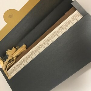 May include: A black and gold invitation envelope with a decorative border and a tassel. The envelope is open to reveal a white card with a brown border.