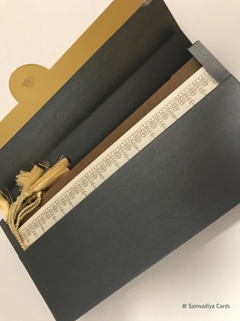 May include: A black and gold invitation envelope with a decorative border and a tassel. The envelope is open to reveal a white card with a brown border.