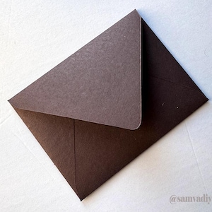 May include: A dark brown paper envelope with a pointed flap and a textured finish. The envelope is positioned diagonally, and the text @samvadiyacards is printed in a small, light-colored font in the bottom right corner of the image.
