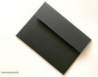 May include: A black envelope with a flap, set against a white background. The envelope is made of a matte paper material. The corner of the envelope is visible, showing the folded construction. The text "@samvadiyacards" is in the bottom left corner.