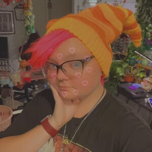 May include: A person with pink hair is wearing a bright orange and yellow striped knit hat. They are wearing a black t-shirt with a graphic print of an anime character.