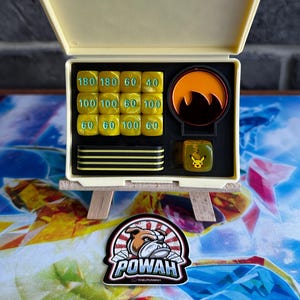 Könnte beinhalten: Eine cremefarbene, offene Box, die einer Malerpalette ähnelt und gelbe Würfel mit schwarzen Zahlen, ein schwarz-oranges Design und einen Pikachu-Würfel enthält. Die Box steht auf einer Miniatur-Holzstaffelei. Unten befindet sich ein Aufkleber mit einer Bulldogge und dem Wort "POWAH".