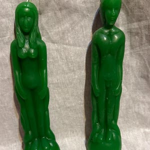 May include: Two green figure candles. One candle depicts a woman with long hair and a flowing dress. The other candle is a male figure. Both candles are standing on a round base. The candles are made of a solid green wax.