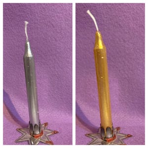 May include: Two taper candles, one silver and one gold, each with a white wick. The candles are set in star-shaped holders with red accents. The background is a solid purple color.