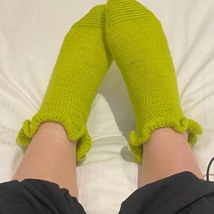 May include: Bright lime green knitted socks with a frilled trim. The socks are made from a textured yarn and are shown against a white backdrop. These socks are a vibrant and stylish accessory.