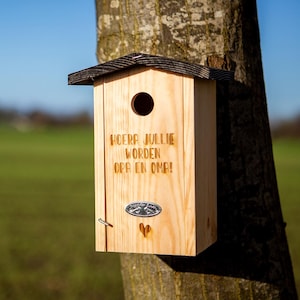 May include: A wooden birdhouse with a dark roof is mounted on a tree trunk. The birdhouse has a round entry hole and engraved text. A small metal emblem and a heart-shaped cutout are visible on the front.