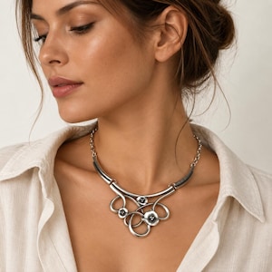 Puede incluir: Un collar de metal plateado con un colgante central compuesto por bucles curvos interconectados y tres pequeños elementos en forma de flor con centros oscuros. La pieza incluye una sección de barra curva lisa y una cadena de eslabones.