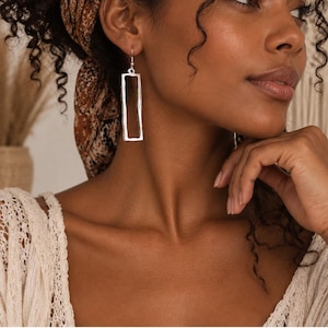 May include: A silver-toned drop earring featuring a rectangular frame with a textured, uneven surface. The earring hangs from a simple hook wire, showcasing a hollow centre and a metallic finish that catches the light with its organic, hand-crafted appearance.