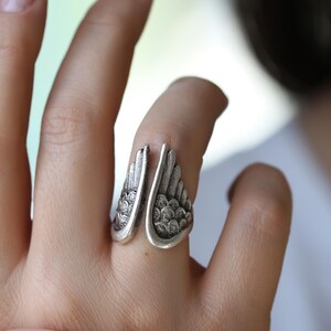 Op de afbeelding: Een zilverkleurige ring met een open ontwerp, met vleugelvormige details. De vleugels van de ring buigen om de vinger, met getextureerde, veerachtige patronen. De ring wordt aan een hand gedragen en toont het ontwerp van de sieraden.