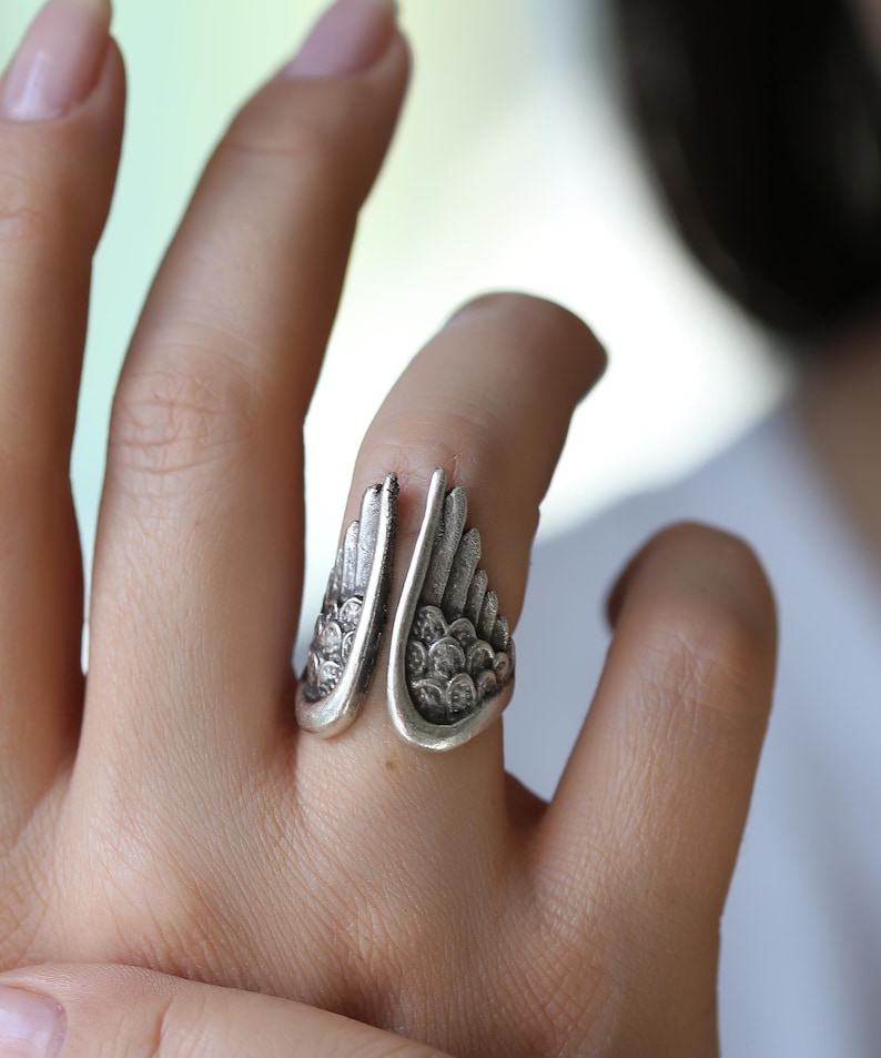 Op de afbeelding: Een zilverkleurige ring met een open ontwerp, met vleugelvormige details. De vleugels van de ring buigen om de vinger, met getextureerde, veerachtige patronen. De ring wordt aan een hand gedragen en toont het ontwerp van de sieraden.