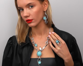 May include: A silver necklace and earrings set featuring turquoise stones. The necklace has a large, round turquoise stone pendant with intricate silver wirework. The earrings are long and dangle, with a smaller turquoise stone at the end.