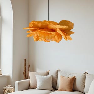 May include: A large, orange paper flower pendant light hangs from the ceiling in a living room. The light fixture is the focal point, with a layered petal design. The room features a neutral color palette with a sectional sofa and a round ottoman.