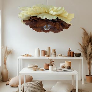 May include: A unique, layered paper flower pendant light fixture. The light has a bulb in the center and is made of cream and brown colored paper. The light hangs above a white table with decorative items.