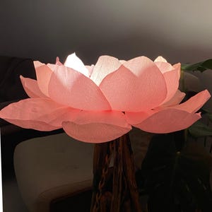 May include: A large, illuminated pink lotus flower lamp. The petals are a soft pink, and the light shines through them, creating a warm glow. The lamp is supported by a brown, textured stem, adding a natural element to the design. The overall effect is elegant and serene.