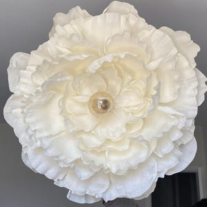 Peut inclure: Une grande fleur en papier crème avec de multiples couches de pétales froissés. Le centre présente un petit luminaire rond de couleur dorée. La fleur est un élément décoratif, peut-être pour des événements ou la décoration intérieure.