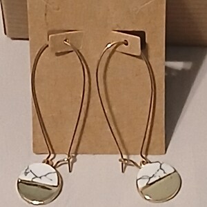 May include: A pair of gold-colored hoop earrings. Each earring features a circular pendant with a white marble design on one half and a light green color on the other half. The earrings are displayed on a brown cardboard backing.