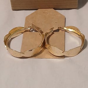 May include: A pair of gold-colored hoop earrings. Each earring has a textured, leaf-like design around the hoop. The earrings are displayed on a brown tag, with a brown cardboard box in the background.