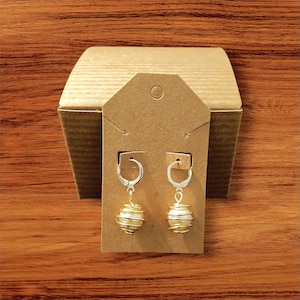 May include: Gold-coloured earrings featuring a spiral wire design around a cream-coloured sphere. The earrings are displayed on a brown earring card with a brown cardboard box in the background. The earrings have a lever back closure.