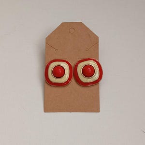 May include: A pair of square-shaped earrings with a red center, surrounded by a cream-colored border and a red outer edge. The earrings have a gold-colored trim and are displayed on a brown tag.