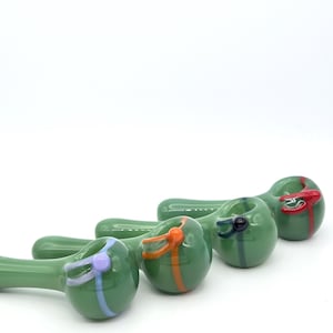 May include: Four green glass pipes arranged in a row, each featuring a distinct colored accent band and a small circular detail. The colors of the accents include light purple, orange, dark blue, and red, contrasting against the smooth glass.