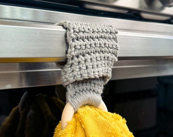 May include: A gray crocheted towel holder with a wooden ring, hanging on a silver oven door handle. A yellow towel is draped over the ring.