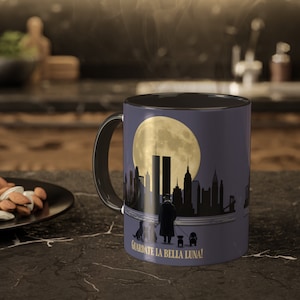 May include: Black coffee mug with a purple background featuring a silhouette of the New York City skyline with the Twin Towers, a full moon, and a man walking four dogs. The text "GUARDATE LA BELLA LUNA!" is printed in gold at the bottom of the mug.