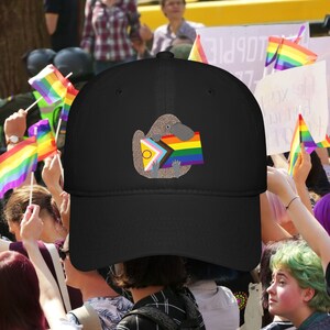 Peut inclure: Casquette de baseball noire avec une image brodée d'un ornithorynque tenant un drapeau arc-en-ciel de la fierté. L'ornithorynque tient le drapeau avec ses pattes avant et a un cercle jaune sur sa poitrine.