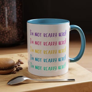 Può includere: Una tazza in ceramica bianca con un bordo e un manico blu. La tazza ha il testo "I'M NOT REALLY HERE!" ripetuto cinque volte in diversi colori.