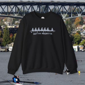 Puede incluir: Sudadera negra con un gráfico blanco de un equipo de remo en un bote. El texto "GOTTA REGATTA" está impreso debajo del gráfico.