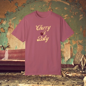 Puede incluir: Una camiseta rosa con el texto "Cherry Baby" en una fuente retro. Una cereza con un tallo está incluida en el texto.