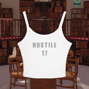 Könnte beinhalten: Weißes Tanktop mit dem Wort "HOSTILE" und der Zahl "17" in Grau aufgedruckt. Das Top hat dünne Träger und einen Rundhalsausschnitt. Der Hintergrund zeigt eine Bibliothek mit Bücherregalen und Holzmöbeln.