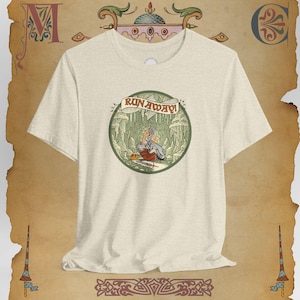 May include: Cream-colored t-shirt featuring a circular illustration with the text "RUN AWAY!". The illustration depicts a rabbit and a person in a forest setting. The shirt has short sleeves and a crew neck.