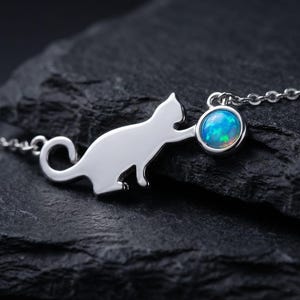 May include: A silver cat pendant necklace with a round, blue opal gemstone. The cat silhouette is detailed, and the necklace chain is delicate. The pendant is displayed on a dark, textured surface.