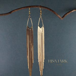 Puede incluir: Un par de pendientes de oro colgantes con un diseño geométrico. Los pendientes están hechos de una cadena de pequeños eslabones de oro que cuelgan de una forma triangular.