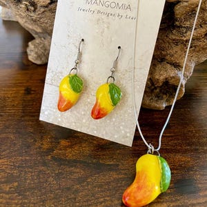 May include: A jewelry set featuring a mango design. The set includes a necklace and a pair of earrings. The mangoes are yellow, orange, and green, with a leaf detail. The jewelry is displayed on a card that reads "MANGOMIA Jewelry Designs by Leai."