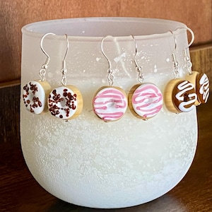 May include: Five pairs of donut earrings. Each pair features a different donut design with white frosting and various toppings, including chocolate sprinkles, pink stripes, and white swirls. The earrings are made of wood and have silver hooks.