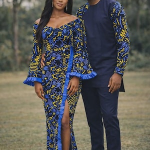 May include: A couple in matching outfits. The woman wears a blue, yellow, and black patterned off-the-shoulder dress with a high slit. The man wears a navy blue shirt with matching patterned sleeves and navy pants.