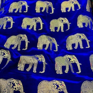 Op de afbeelding: Koningsblauwe stof met een herhaald patroon van gouden olifanten. De olifanten zijn gedetailleerd met gouden contouren en ingewikkelde ontwerpen, waardoor een luxueus en opvallend textiel ontstaat.