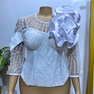 Puede incluir: Top blanco con cuentas y un gran volante blanco en un hombro. El top tiene un corpiño ajustado y mangas largas transparentes.