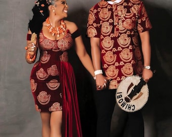 May include: A couple in traditional Nigerian attire. The woman is wearing a maroon dress with gold embroidery and a long maroon train. The man is wearing a maroon shirt with gold embroidery and a matching hat. The man is holding a wooden fan with the word "CHIMOBI" written on it.