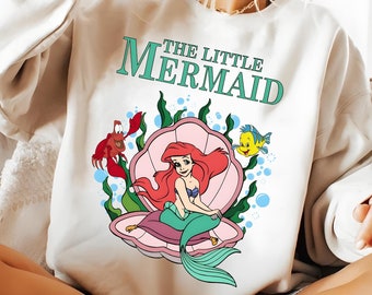 May include: A white sweatshirt with a graphic of Ariel, the Little Mermaid, sitting on a seashell. The text "THE LITTLE MERMAID" is printed above the graphic.