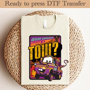 May include: A beige t-shirt with a cartoon tow truck graphic. The tow truck is purple and has a smiling face. The text on the graphic says "Gotcha Covered Need a Tow?"