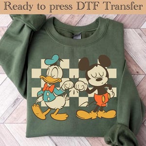 May include: Green sweatshirt with a vintage-style graphic of Mickey Mouse and Donald Duck giving each other a high five. The graphic is on a checkered background.