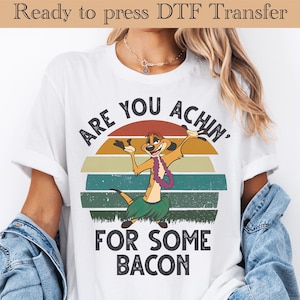 May include: White t-shirt with a graphic of a cartoon meerkat wearing a lei and grass skirt, set against a sunset design. The text reads "ARE YOU ACHIN' FOR SOME BACON".